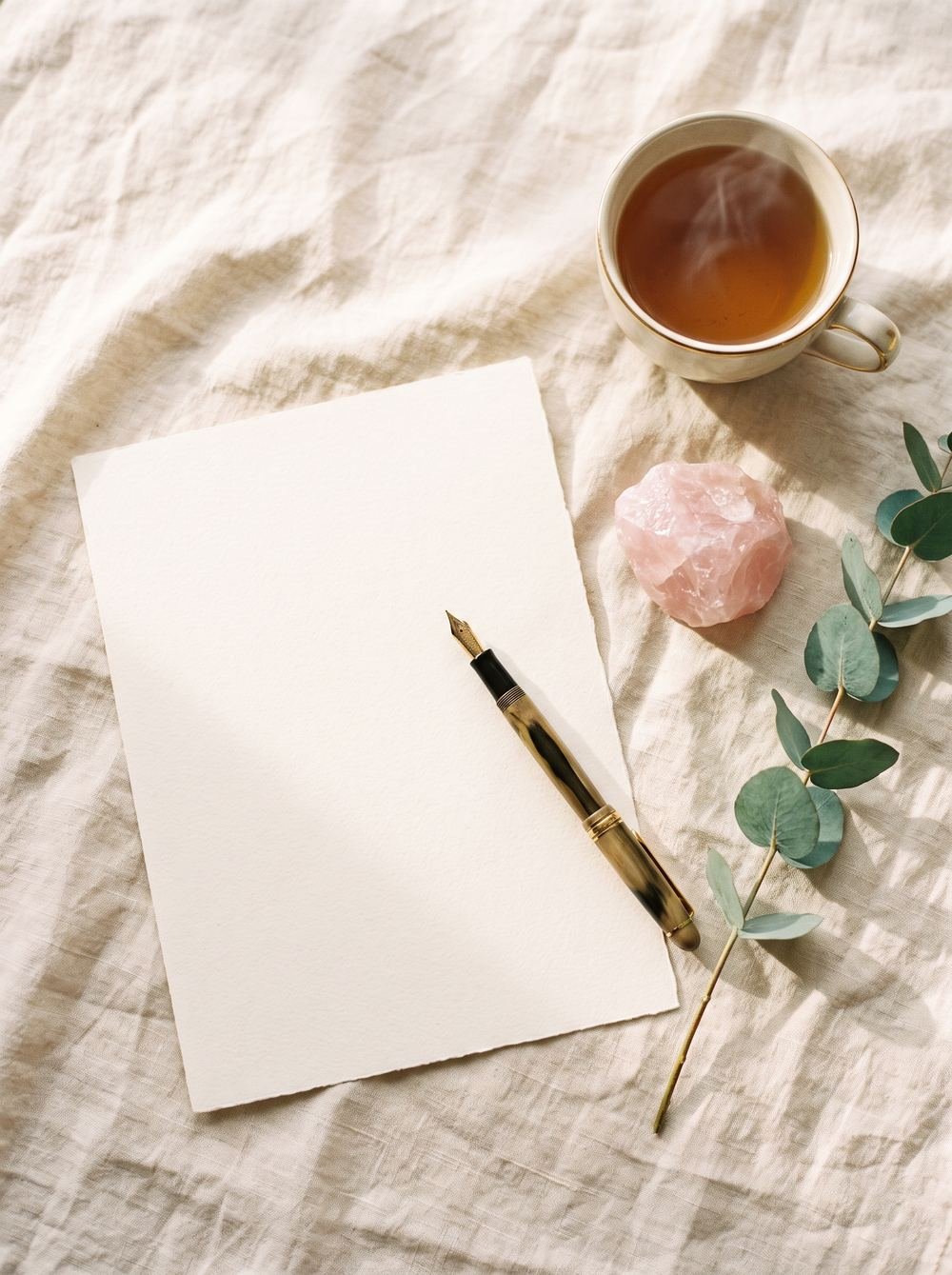 Cream writing paper with a fountain pen, a rose-quartz stone, eucalyptus and a cup of tea on linen