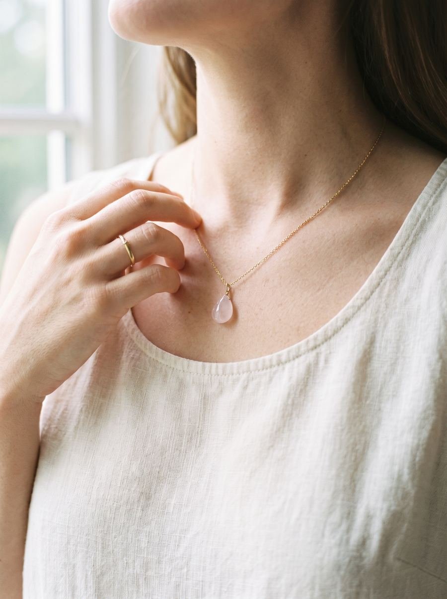 A fine gold necklace with a small rose-quartz pendant, worn over a linen top in soft morning light
